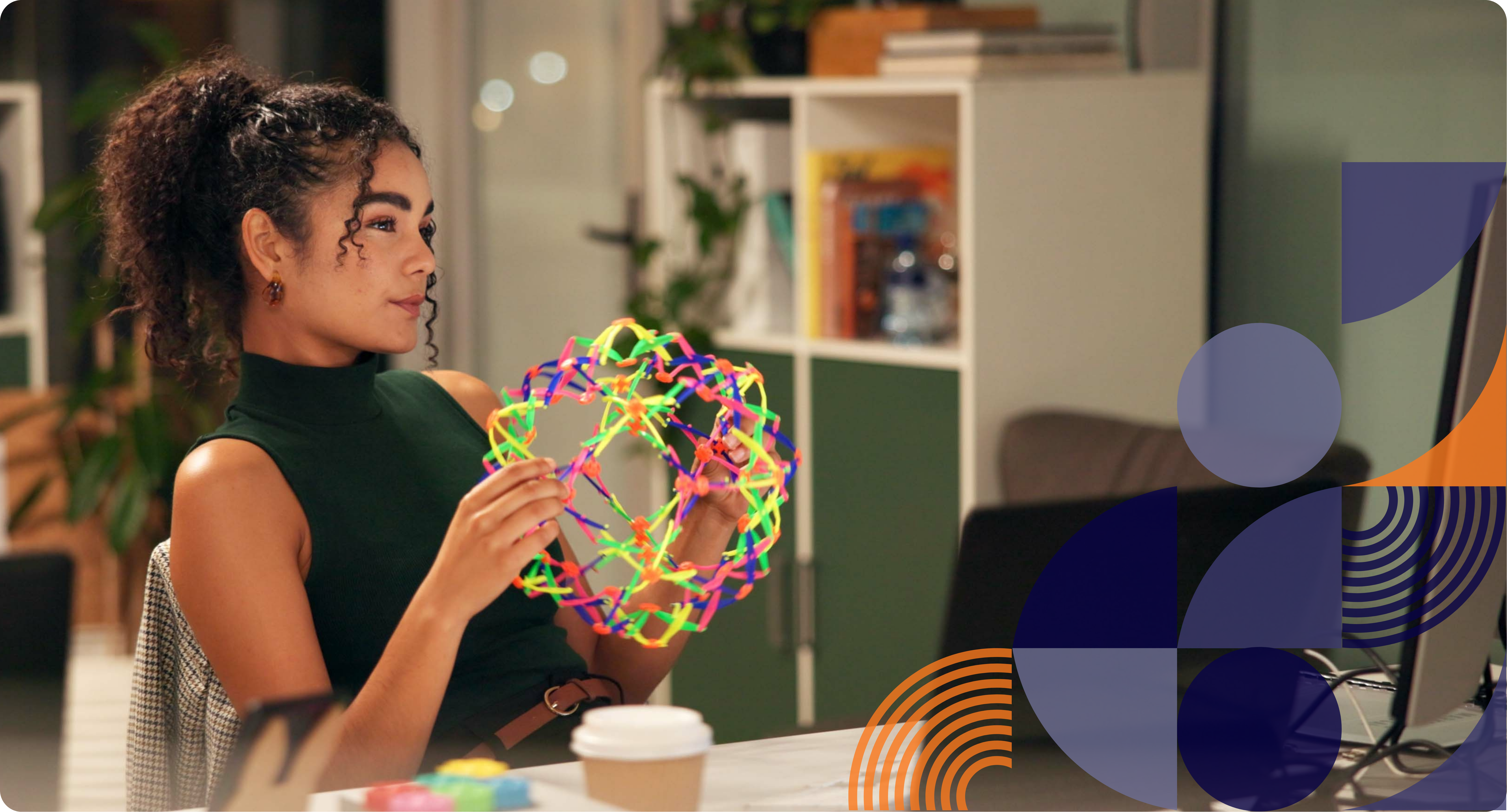 A person with curly hair tied back sits at a desk, holding a colorful collapsible geometric sphere fidget. They wear a sleeveless dark green top, and their posture suggests curiosity or engagement with their laptop. In the background, a bookshelf filled with books and personal items is visible, along with a computer monitor on the desk.